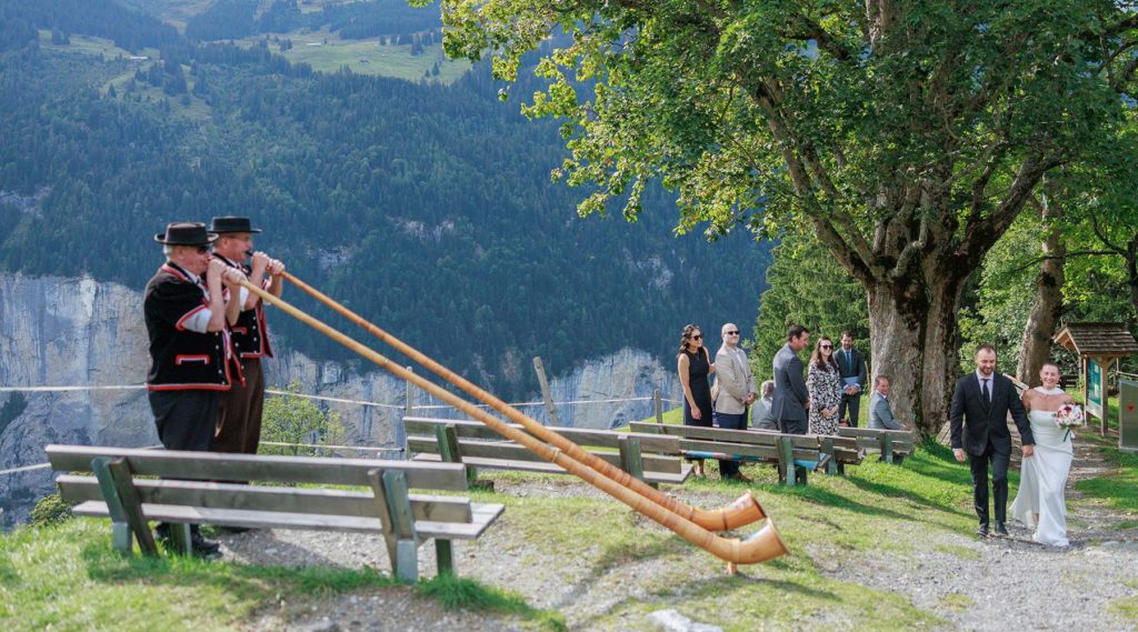 Two men in traditional alpine attire play alphorns at a Swiss wedding ceremony overlooking a mountain valley, as the couple walks toward guests seated on wooden benches under a large tree.