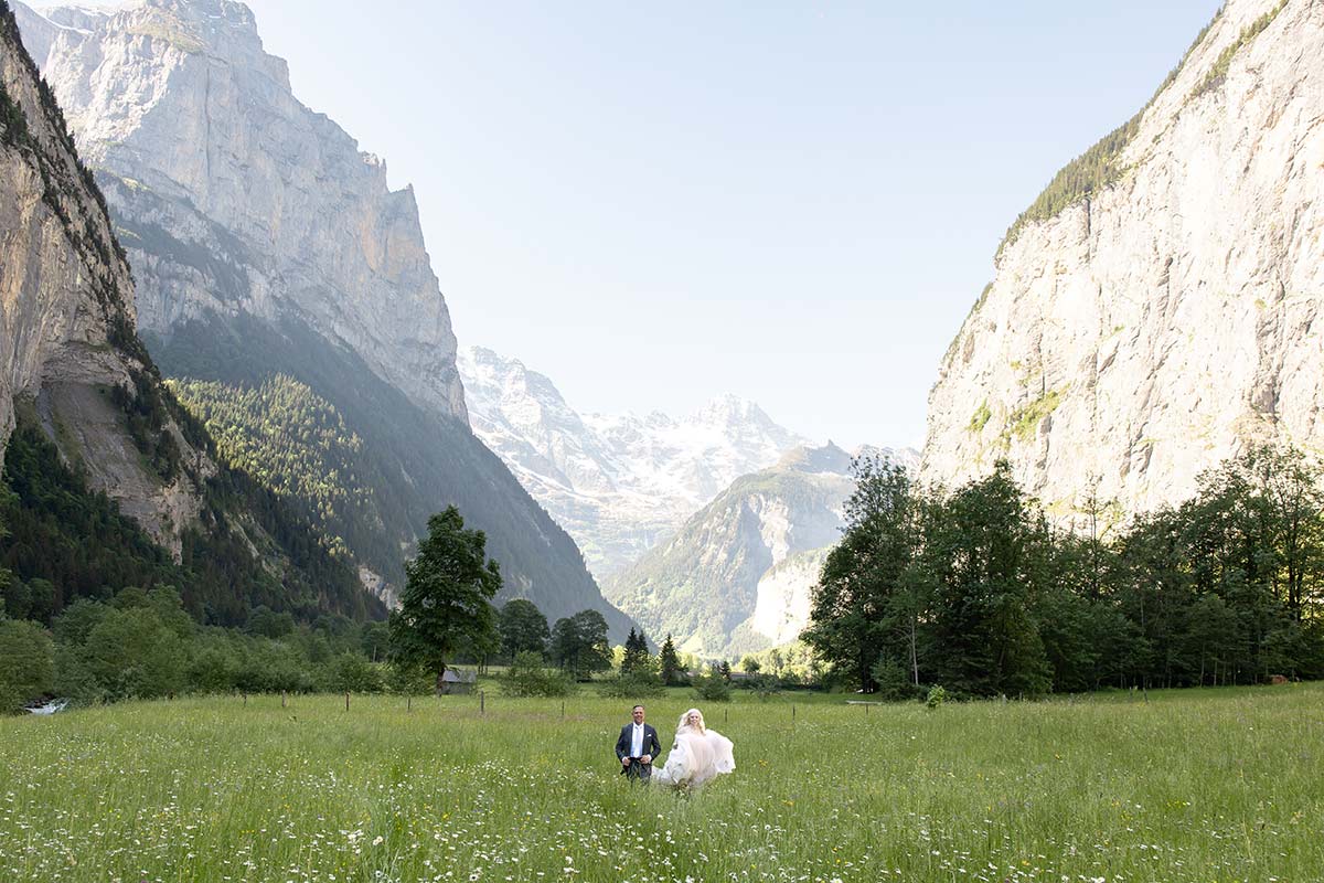 A bride and groom walk through a wildflower meadow in Switzerland surrounded by high mountain cliffs.