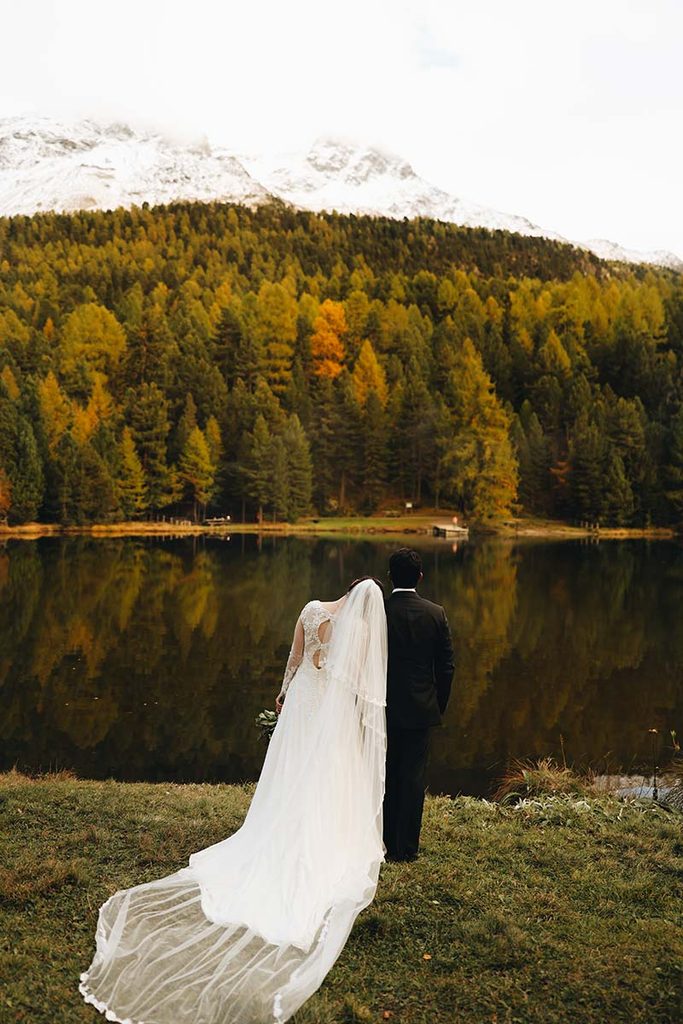 A bride and groom stand by a lake in Switzerland, looking toward a forest and snow-capped mountains.