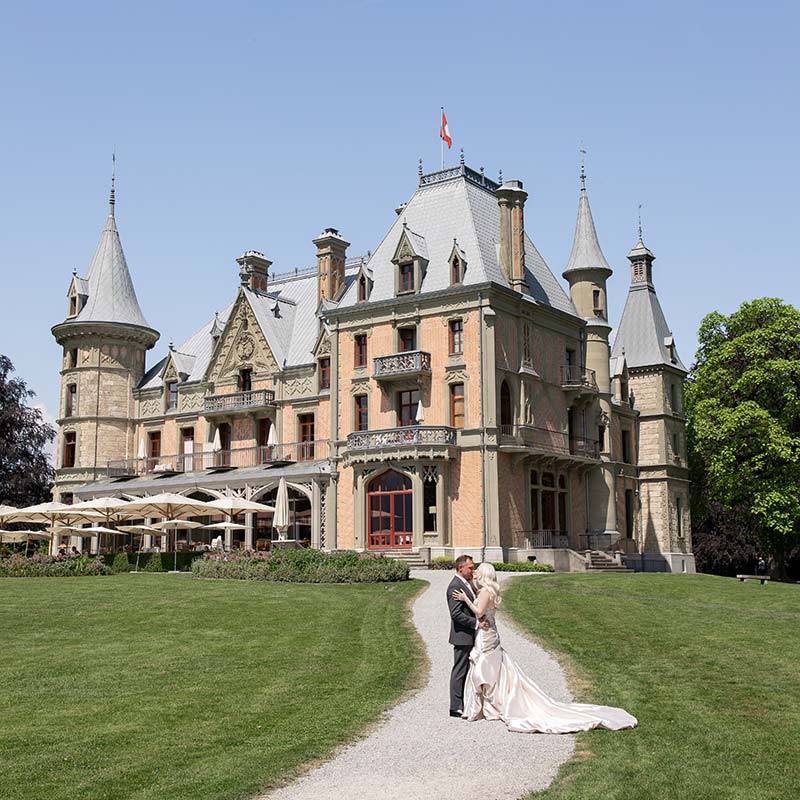 A couple stands on a gravel path in front of a large stone chateau with multiple towers during a tour of Switzerland.