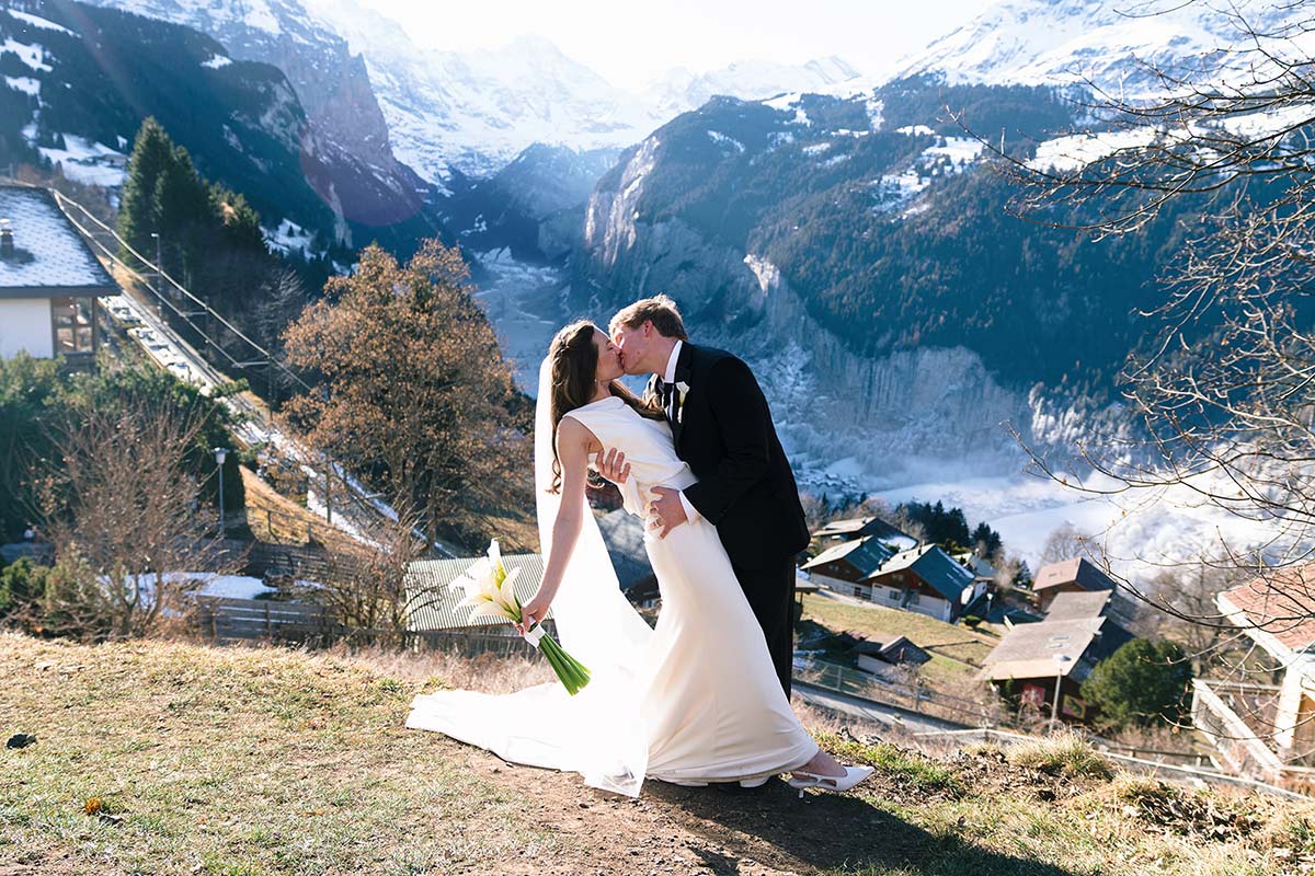 A bride and groom kiss on a Swiss hillside overlooking a snowy mountain valley.