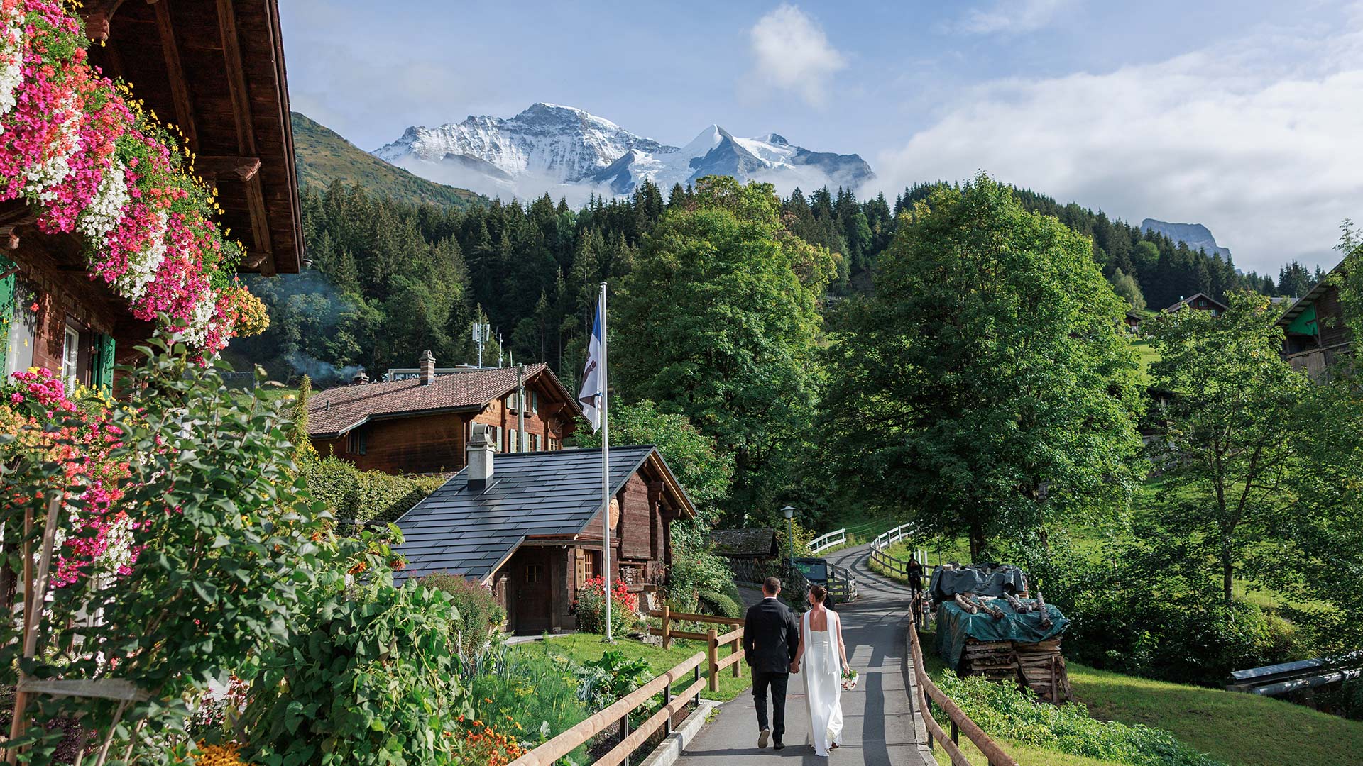 A bride and groom walk hand-in-hand along a path in a Swiss village with snow-capped mountains in the background.