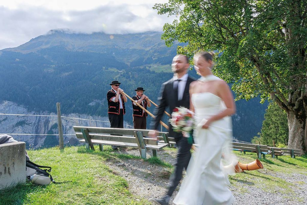 A bride and groom walk past two men playing alphorns in a Swiss mountain landscape during a wedding videography session.