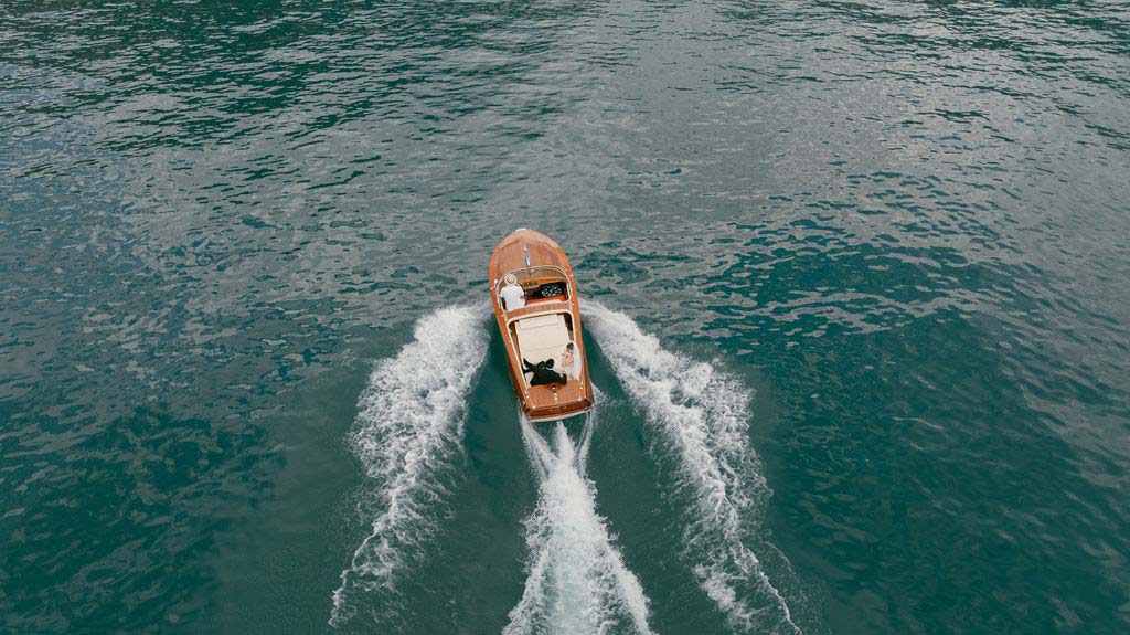 Aerial view of a wooden boat with two people on board moving through blue water, leaving a white wake, captured by a Switzerland wedding videographer.