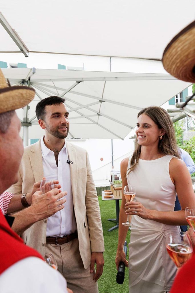 A man in a tan suit and a woman in a white dress talking while holding drinks at a Switzerland wedding videography gathering.