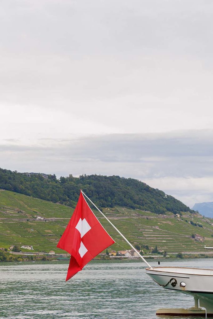 The Swiss flag flies on the bow of a wedding videography boat on a lake with terraced hills in the background.