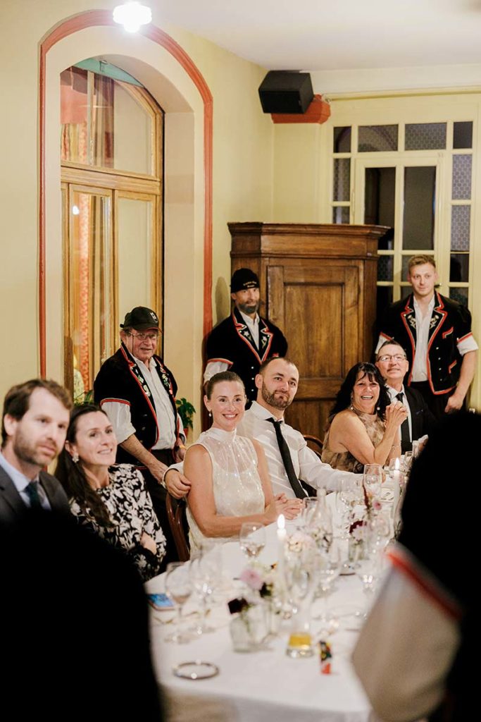 A wedding reception table with guests seated, including a bride and groom, while servers in traditional Swiss outfits stand behind them.