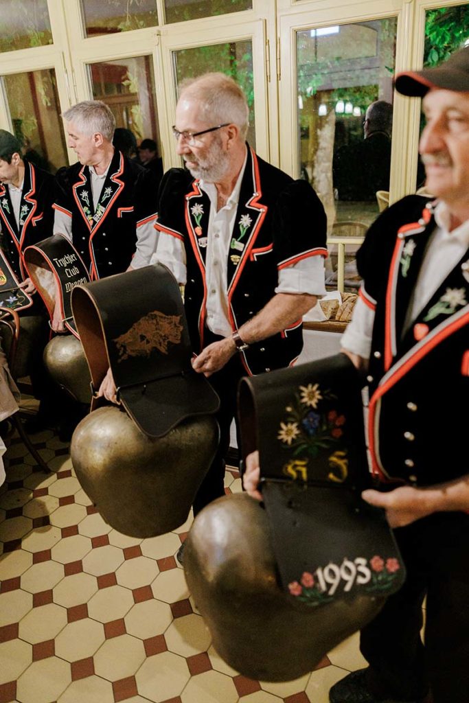 Three men in traditional black Swiss outfits holding large metal cowbells with decorative leather straps.