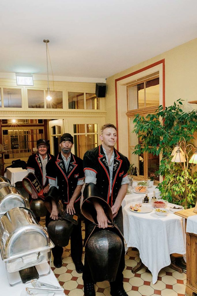 Three men wearing traditional Swiss outfits and hats stand in a dining room holding large brass cowbells.