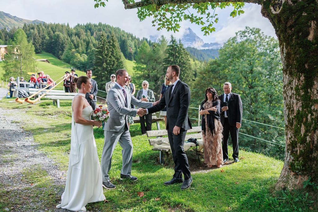 A bride and groom shake hands outdoors in a mountain setting captured by a Switzerland wedding videographer, with wedding guests nearby.